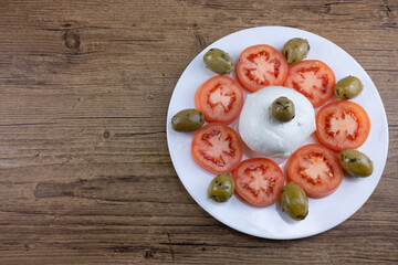 The meal on a white plate and wooden table consists of sliced tomatoes, pitted olives and mozzarella