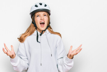 Beautiful young caucasian girl wearing bike helmet crazy and mad shouting and yelling with aggressive expression and arms raised. frustration concept.