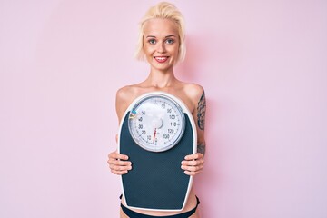 Young blonde woman with tattoo standing shirtless holding weighing machine smiling with a happy and cool smile on face. showing teeth.