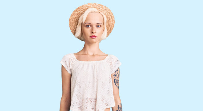 Young blonde woman with tattoo wearing summer hat relaxed with serious expression on face. simple and natural looking at the camera.