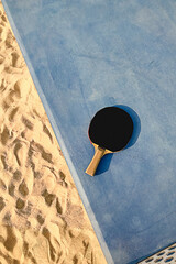 Black tennis racquet on a blue table outside at the beach sand on a sunny day top view. Outdoor sport concept.