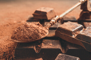 Brown cocoa powder in the spoon and chopped chocolate cubes, on dark background