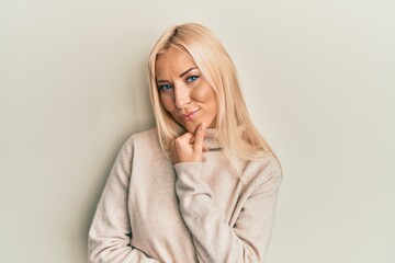 Young blonde woman wearing casual winter turtleneck sweater smiling looking confident at the camera with crossed arms and hand on chin. thinking positive.