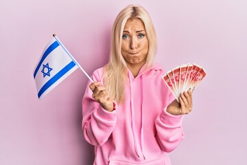 Young blonde woman holding israel flag and shekels banknotes puffing cheeks with funny face. mouth inflated with air, catching air.