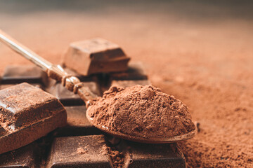 Brown cocoa powder in the spoon and chopped chocolate cubes, on dark background with copy space