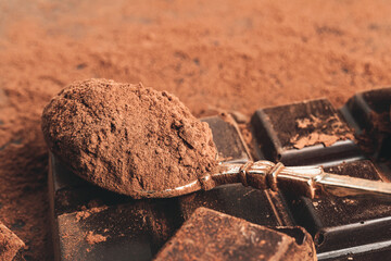 Brown cocoa powder in the spoon and chopped chocolate cubes, on dark background
