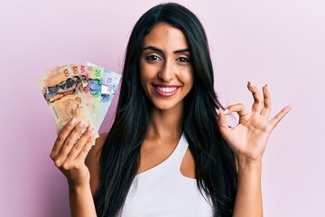 Beautiful hispanic woman holding canadian dollars doing ok sign with fingers, smiling friendly gesturing excellent symbol