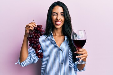 Beautiful hispanic woman holding branch of fresh grapes and red wine sticking tongue out happy with...