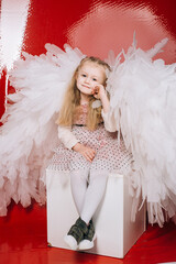 little girl 4 years old in white angel wings on a red latex background in the studio for valentine's day
