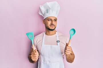 Young arab man wearing professional cook apron and hat holding spoon smiling looking to the side...
