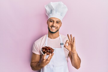 Young arab man wearing professional cook uniform holding bowl with dates doing ok sign with fingers, smiling friendly gesturing excellent symbol