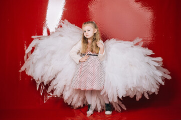 little girl 4 years old in white angel wings on a red latex background in the studio for valentine's day