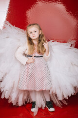little girl 4 years old in white angel wings on a red latex background in the studio for valentine's day
