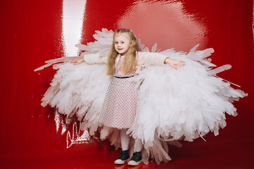 little girl 4 years old in white angel wings on a red latex background in the studio for valentine's day