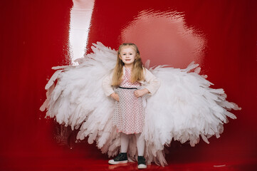 little girl 4 years old in white angel wings on a red latex background in the studio for valentine's day