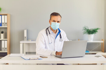 Portrait of a serious doctor in a protective mask and medical gown sitting at his desk with a laptop and looking at the camera. Concept of healthcare and modern technology.