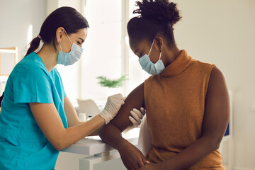 Young African American woman getting flu shot during seasonal vaccination campaign. Doctor or nurse in medical face mask cleans skin on patient's arm before injecting modern Covid 19 antiviral vaccine - obrazy, fototapety, plakaty