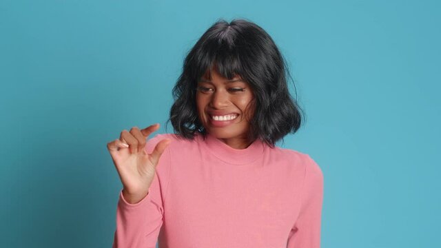 Young brunette Asian woman with dark skin shows very tiny object shapes small item needs some more dressed in casual clothing poses against blue background. Minimum size. I need not much