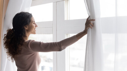 Smiling young dreamy woman opening curtains, looking outside window in morning. Sincere millennial caucasian lady breathing fresh air, starting new day or enjoying peaceful weekend alone at home.