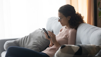 Naklejka na ściany i meble Relaxed young woman in glasses sitting on cozy sofa, involved in chatting in social networks using mobile application, web surfing information, shopping in online store or ordering delivery food.