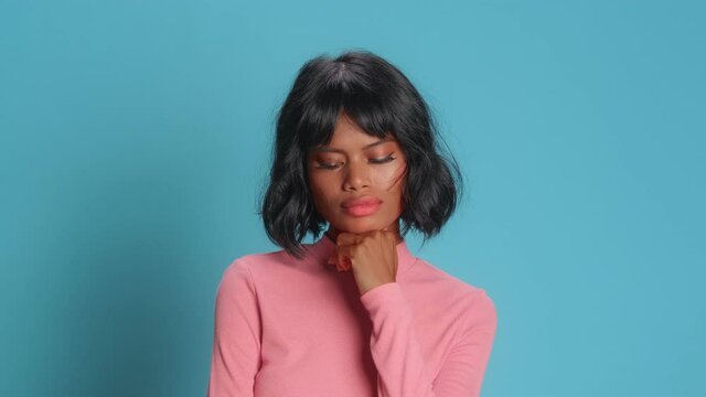 Determined thoughtful young woman with dark skin wavy bushy hair holds chin and looks away pensively tries to make decision dressed casually poses against blue background. Let me think concept