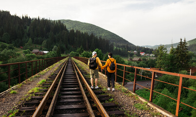 Loving couple of tourists in casual clothes walking holding hands on the railway bridge against the backdrop of a mountain landscape.