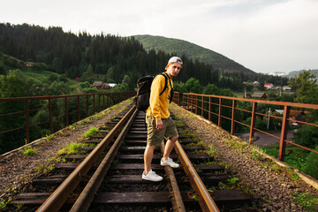 Fototapeta na wymiar A guy in an orange sweater stands on a track on a railway bridge in the mountains and looks at the camera. young male tourist walks on a viaduct in the mountains