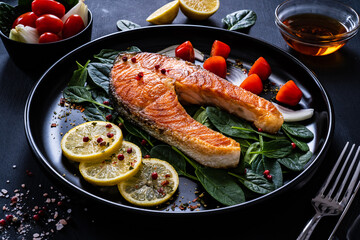 Fried salmon steak with spinach, lemon and cherry tomatoes  served on black plate on wooden table

