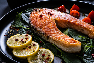 Fried salmon steak with spinach, lemon and cherry tomatoes  served on black plate on wooden table
