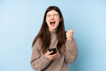 Young Ukrainian teenager girl wearing a sweater over isolated blue background with phone in victory position