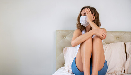 Quarantine at home concept. Unhappy young woman wear casual cloth and protective face mask worried something while sitting alone on white bed in the room.