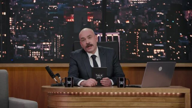 ZOOM IN Late-night talk show host sitting behind his table and performing his monologue, looking into camera. TV broadcast style show. Shot with RED cinema camera