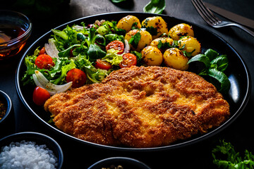 Breaded fried pork chop, fried potatoes and fresh vegetables on black table