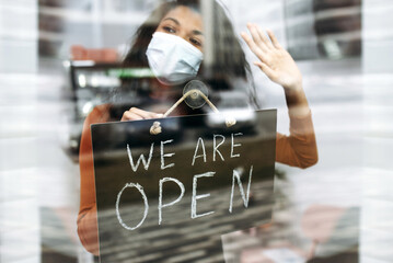 African American small business owner in medical mask holds a signboard WE ARE OPEN at the entrance to a restaurant, bar, cafe or shop, and waiting for visitors