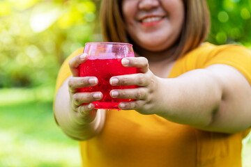 Eat a lot. Not good for health. Overweight obesity Unhealthy concept. Close up hands of happy smile fat young woman holding a glass of red nectar with ice soda while standing in the garden.
