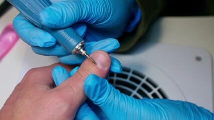 Manicurist in blue gloves removes dry skin around nail with machine nailfile on man's hand. Close up view.