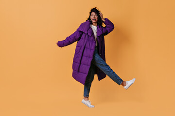 Amazed mixed race girl in down jacket standing on one leg. Full length view of dancing curly woman in jeans.