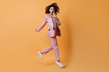 Fototapeta na wymiar Full length view of shocked curly woman. Amazed lady in pink suit jumping on yellow background.