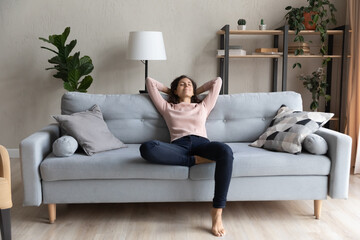 Full length peaceful happy young woman relaxing on comfortable sofa, breathing fresh air, enjoying mindful lazy moment indoors. Smiling millennial caucasian lady meditating alone in living room.