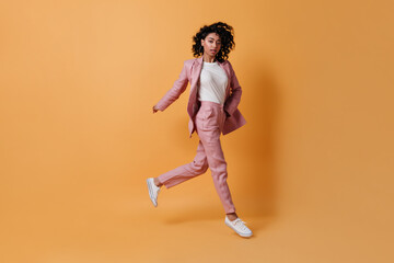 Fototapeta na wymiar Full length view of running curly woman. Adorable girl in pink suit jumping on yellow background.