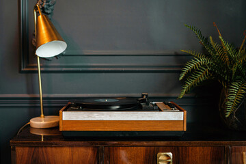 vintage vinyl record player in a stylish interior. a golden candle and green house plants on a vintage wooden table. designer accessories are a table lamp and a book holder. - Powered by Adobe