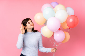 Young Ukrainian teenager girl holding lots of balloons over isolated pink background listening something