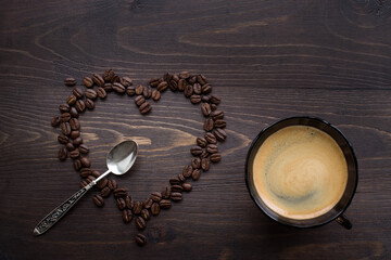 Still life of roasted coffee grains in the shape of a heart and love symbol on a brown wooden background with a cup of coffee and a silver spoon