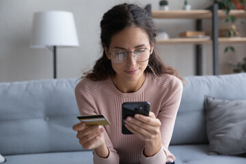 Head shot happy young female client in glasses looking at smartphone screen, entering payment information from credit card in mobile shopping application, enjoying purchasing goods or services online.