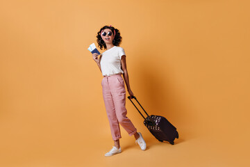 Full length view of female traveler in sunglasses. Studio shot of mixed race curly girl with suitcase.
