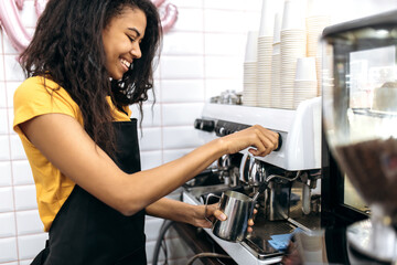 Smiling african american female barista wearing uniform makes coffee for a coffee shop visitor. Cafe, small business concept