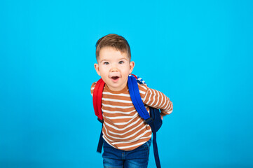 Portrait of surprised happy three year old preschooler with a backpack on blue background. For the first time to school. The concept of developing activities.