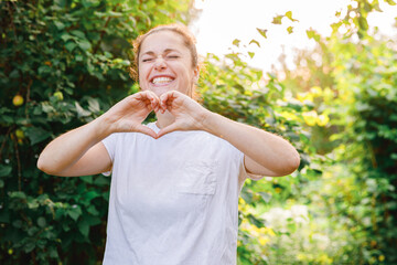 Fototapeta premium Young woman smiling outdoor. Beautiful brunete girl showing heart sign with hands on park or garden green background. Free happy woman at summertime. Freedom happiness carefree happy people concept