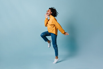 Full length view of woman jumping on blue background. Studio shot of excited lady in jeans.