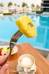 A piece of yellow pineapple on a silver fork at a breakfast time next to the pool. Eating healthy during travel and vacation concept.